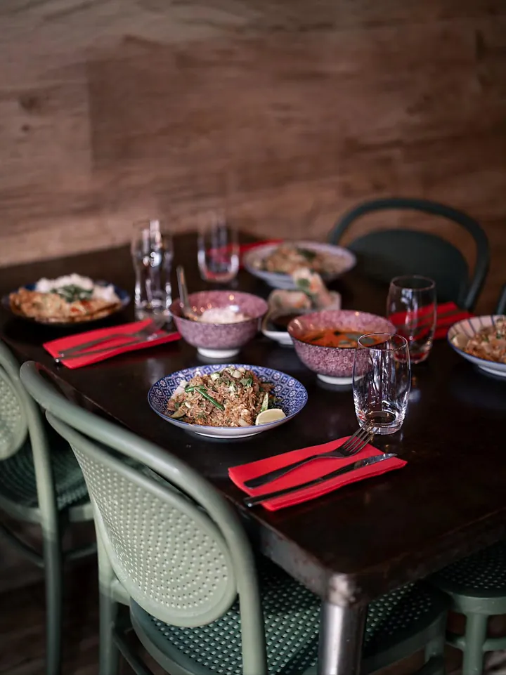 Une table sombre dressée avec des plats en bols, des serviettes rouges et des chaises vert sauge.