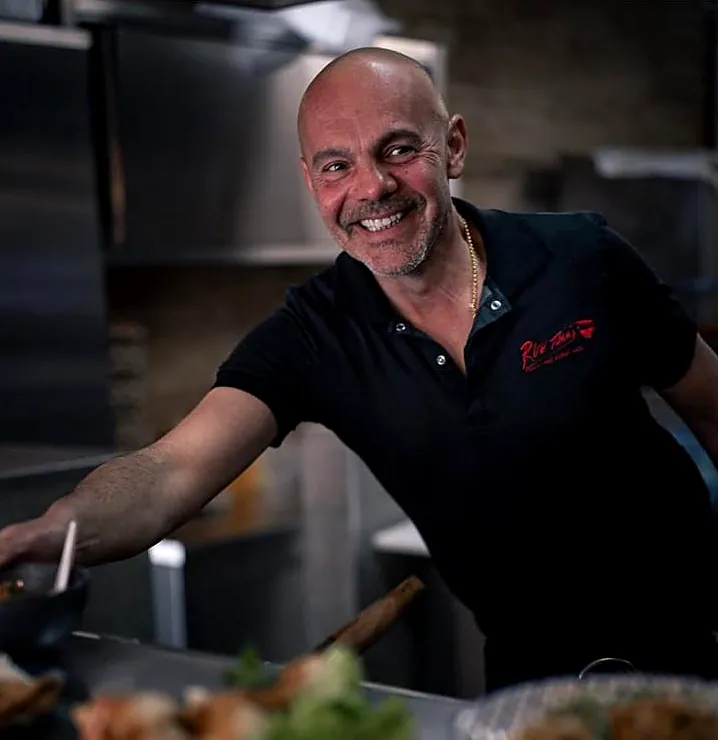 Un cuisinier de Rue Thaï, souriant, en polo noir au logo, devant la cuisine ouverte.