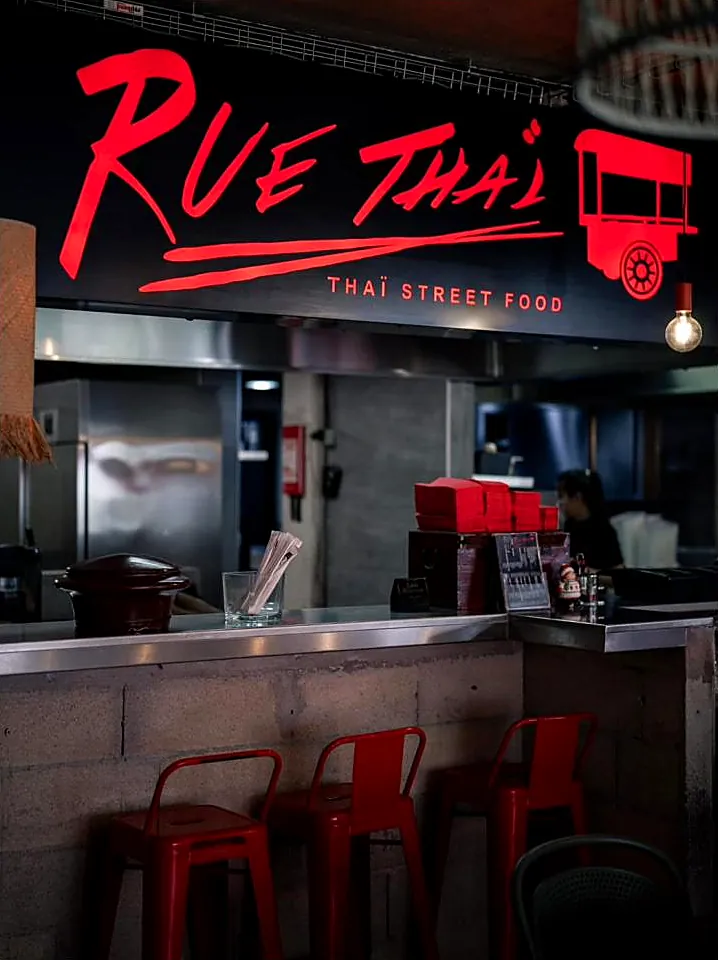L'enseigne rouge lumineuse Rue Thaï au-dessus du comptoir en parpaings et inox, tabourets rouges.