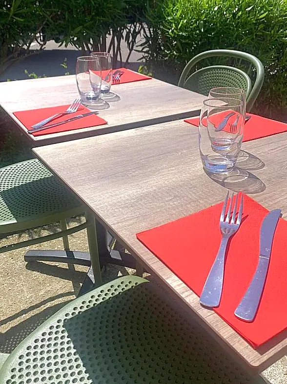 La terrasse au soleil : tables en bois, chaises vert sauge, serviettes rouges, haie de lauriers.